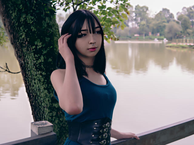 Vickmon met zwart haar, een blauwe jurk, een zwarte korset, een choker en een neusring, met haar hand in haar haar, staand bij een boom, water op de achtergrond, kijkend naar de camera, schattig, sexy..