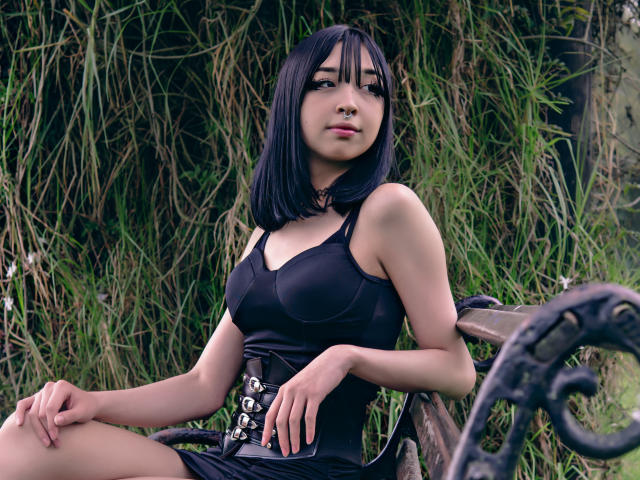 Vickmon met een zwart bob kapsel, gekleed in een zwarte jurk en corset, zittend op een metalen bank, handen op haar dijen, groen gras op de achtergrond, kijkt iets naar beneden, neusring, subtiele make-up..