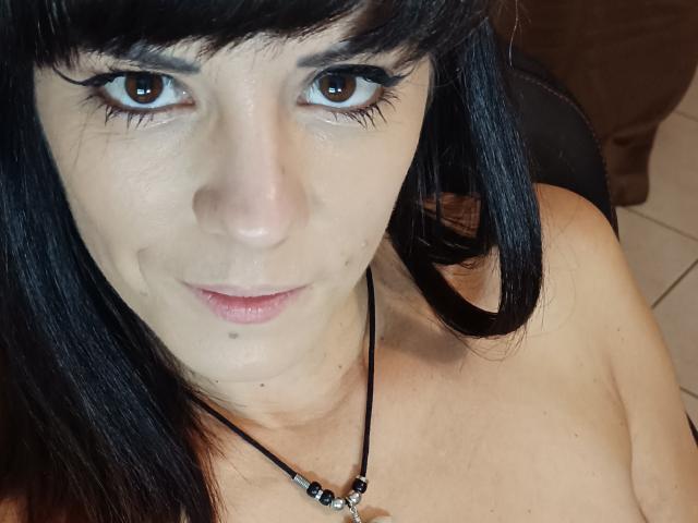 OrianaLaFrancaise has black hair, brown eyes, and a slight smile. She's topless, wearing a black necklace with beads, and has a small mole on her chin.