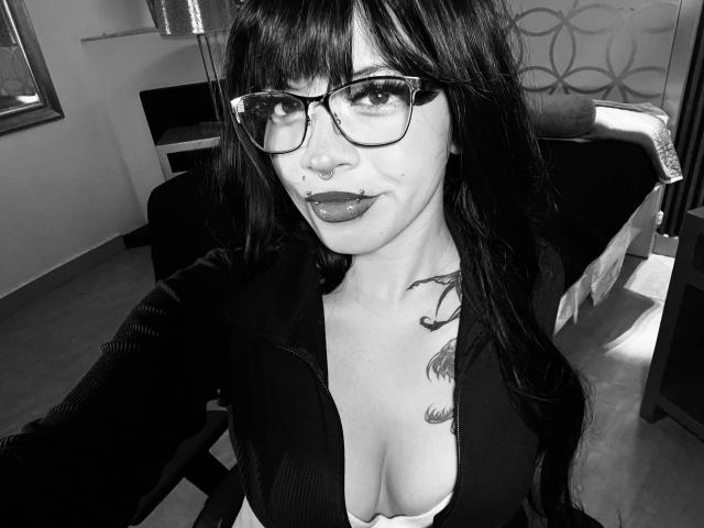 Black-and-white selfie of BabeSix with long dark hair, glasses, nose ring, and dark lipstick, wearing a low-cut top showing her tits, and a tattoo on her right chest.