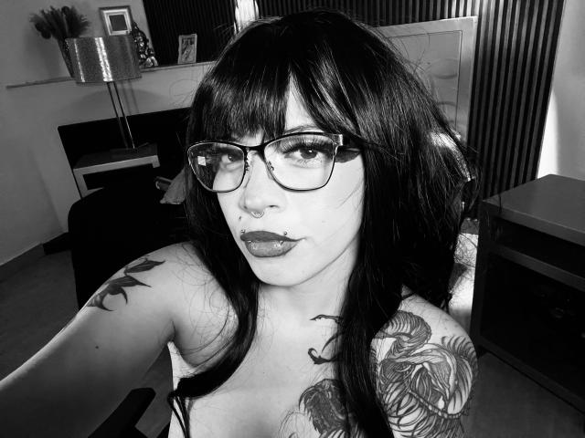 Black-and-white selfie of BabeSix with glasses, dark hair, and tattoos on her shoulder and chest. She's looking at the camera with a nose ring and plump lips.