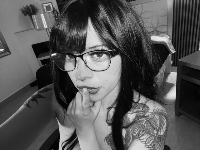 Black-and-white selfie of BabeSix with long dark hair, glasses, nose ring, lip ring, finger in mouth, showing tattooed shoulder, and small tits. BabeSix looks sexy and nerdy.