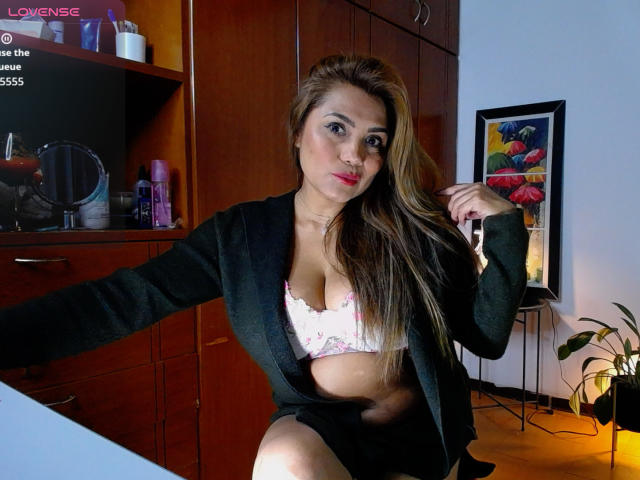 LarisMilfHot with long brown hair, red lipstick, green cardigan, pink floral bra, and black skirt, showing her tits, holding hair, sitting in a wooden room, smiling.