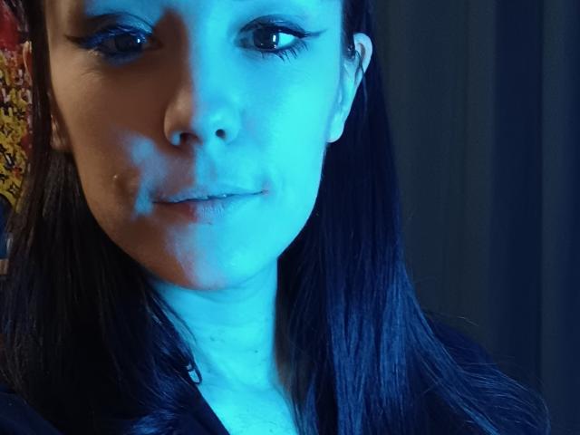OrianaLaFrancaise ha una pelle leggermente azzurrina, capelli neri lisci e un trucco per gli occhi molto leggero, con un piccolo brufolo sulla guancia destra e un sorriso appena accennato, con la bocca chiusa..