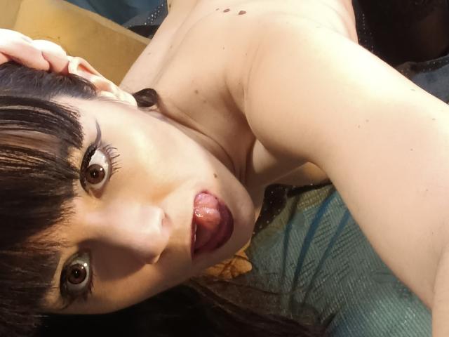 OrianaLaFrancaise with wide eyes, tongue out, and hand in dark hair, topless, showing small moles on her fair skin, and dark makeup. Her tits are partially visible.