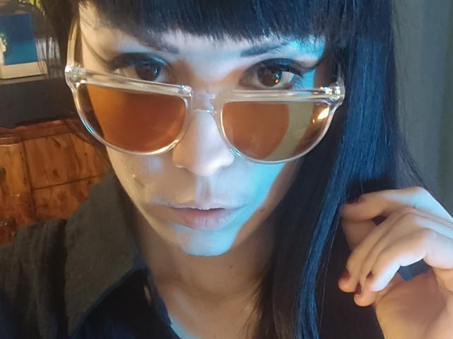 OrianaLaFrancaise with dark straight hair, bangs, and clear aviator sunglasses, blue light on her face, hand near her chin, looking into camera. Her ass is not visible.
