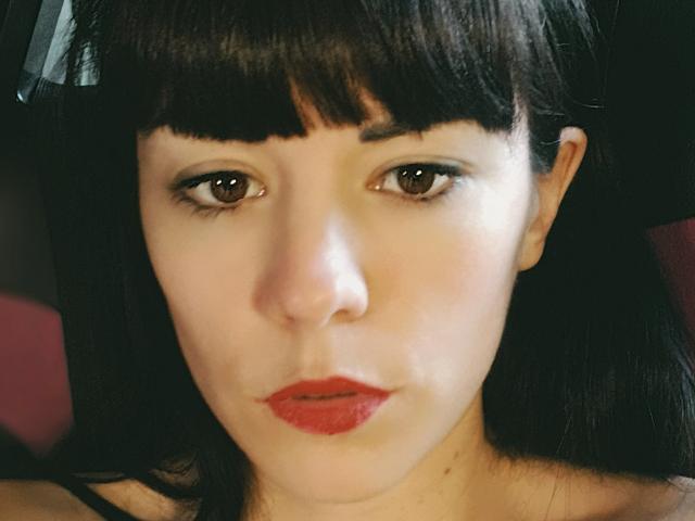 OrianaLaFrancaise a les yeux marron foncé, du rouge à lèvres, et une frange noire, avec une peau pâle et des cheveux noirs lisses. Son expression est sérieuse, et elle regarde directement l'appareil photo..