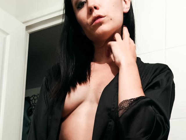 OrianaLaFrancaise, con i capelli neri, indossa un abito di seta nera aperto, che mostra i suoi piccoli seni e una manica con pizzo, una mano sul collo, guarda la fotocamera, sfondo con piastrelle bianche..