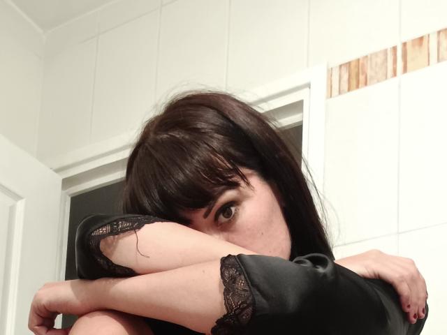 OrianaLaFrancaise con capelli castano scuro, manica in pizzo nero, braccia incrociate sul petto, un occhio che spunta, che guarda la fotocamera, in un bagno con piastrelle bianche..