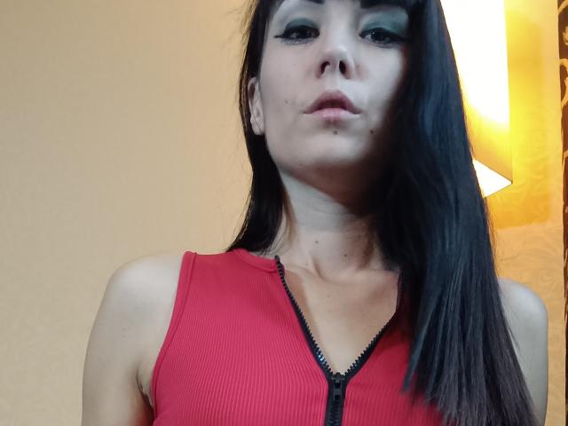 Oriana, una ragazza con lunghi capelli neri, trucco verde sugli occhi, labbra piene e una maglia rossa a coste con zip, che lascia scoperte le spalle e parte del petto. Luce intensa a destra..