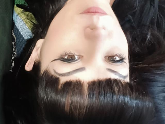Das Gesicht von OrianaLaFrancaise, umgedreht, zeigt dunkelbraune Augen mit dicker schwarzer Eyeliner, schwarzen Augenbrauen und geraden schwarzen Pony, und liegt auf einer dunklen Oberfläche..