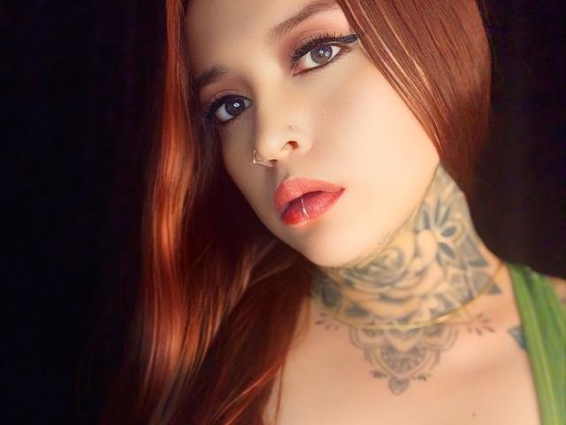 ZoeCaloway with red hair, blue eyes, nose ring, lip piercing, red lipstick, green top, tattooed neck, looking at camera, light skin, makeup, soft light, slightly parted lips.