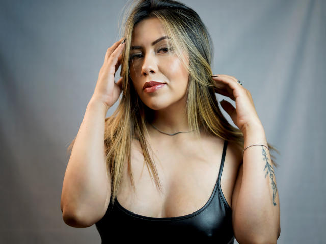 EmilyDurand with blonde hair, tan skin, and a tattoo on her right arm, wearing a black leather top, hands in her hair, looking at the camera with a slight smile.