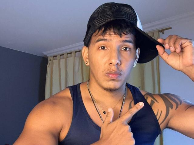 YaoiBoy, um sujeito musculoso com pele morena e tatuagens pretas, usando um boné preto, uma regata azul marinho e brincos, fazendo um gesto de "silêncio" com o dedo..