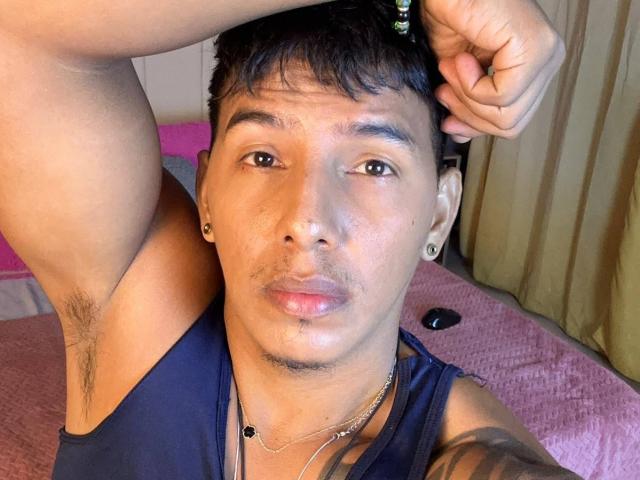 YaoiBoy com cabelo escuro, olhos castanhos e um leve bigode, segurando a cabeça, mostrando um braço tatuado, pelos nas axilas e vestindo uma camisa preta..