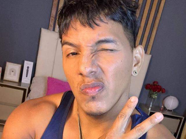 YaoiBoy, um cara latino fofo com cabelo preto curto, está fazendo uma careta engraçada, com os lábios franzidos e um olho fechado. Ele está vestindo uma camiseta azul de alças..