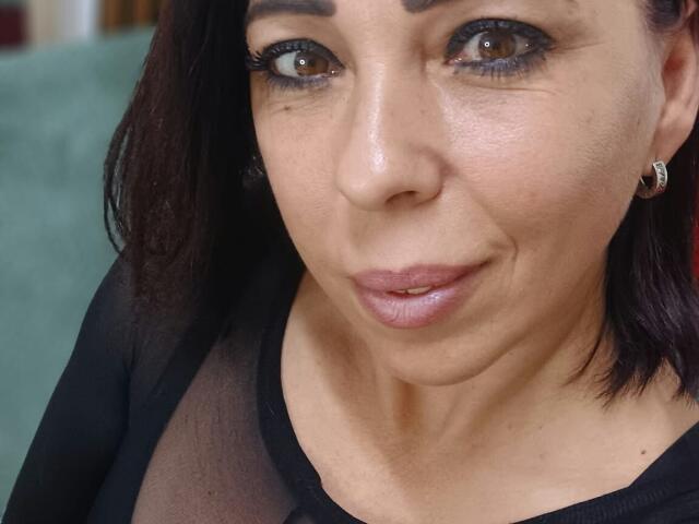 LauraSin's face, showing brown eyes, black eyebrows, medium-length dark hair, wearing a sheer black top, small hoop earrings, slightly parted pinkish lips, and a subtle smile.