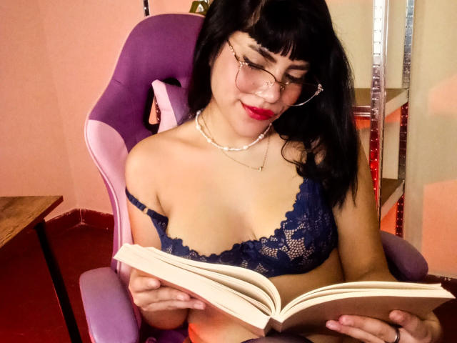 AfroditaMooreX avec des cheveux noirs, des lunettes, du rouge à lèvres rouge, un soutien-gorge en dentelle bleu, lisant un livre, assise sur une chaise violette, un collier de perles, mignonne, sexy, petite poitrine, peau claire, détendue, à l'intérieur..
