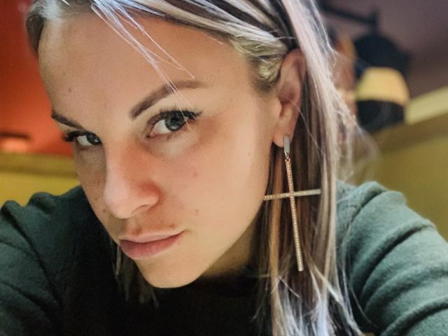 BelleLisaG has light blonde hair, green eyes, and wears a green sweater with cross-shaped earrings. Her face is slightly flushed, and she's giving a serious, subtle pout.