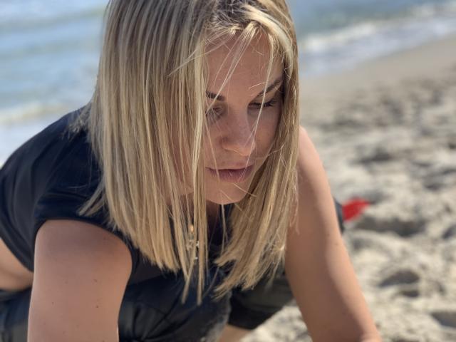 BelleLisaG with blonde hair, black shirt, and freckles, leaning on sandy beach, eyes partially closed, hair in face, sunlit, looking down, red object blurred in background.