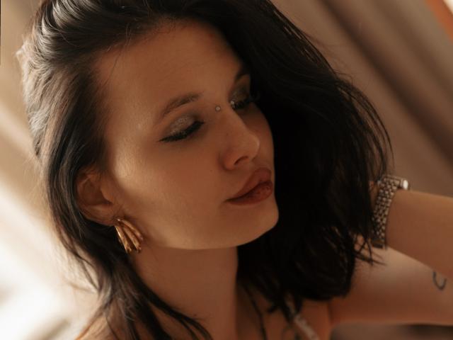 AlicePertim has dark brown hair, a gold earring, and a small beauty mark above her right eyebrow. She's looking down with smoky eye makeup and red lipstick.
