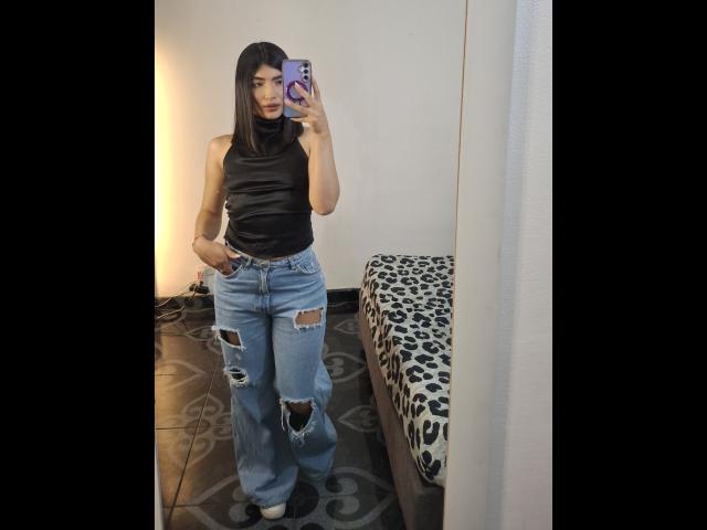 PaulethJoens draagt een zwart topje zonder mouwen, versleten blauwe jeans en heeft haar hand in haar zak, terwijl ze een spiegelselfie maakt. Haar lange bruine haar en rode lippenstift vallen op. Ze houdt een paarse telefoon vast..
