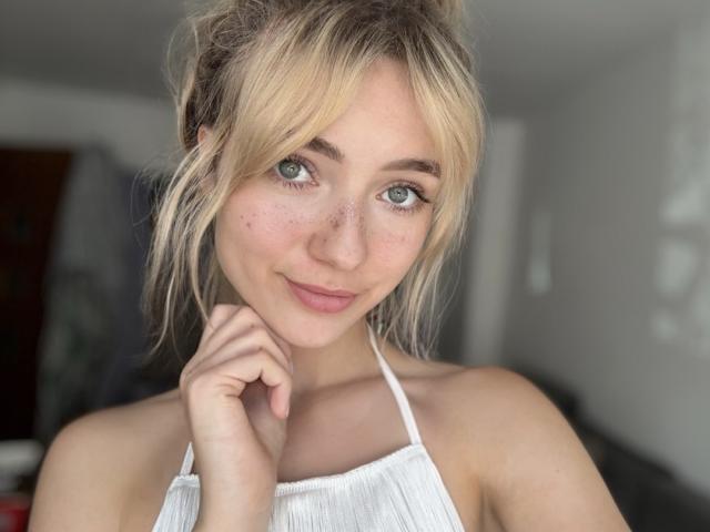 LuciaWild with blonde hair, blue eyes, freckles, and a white halter top, smiling and touching her chin, showing her tits. Blurred background, soft light.