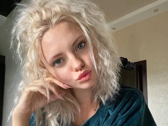 CrystalHope with curly blonde hair, blue eyes, and pouty pink lips, wearing a teal shirt, resting her chin on her hand, looking directly at the camera.