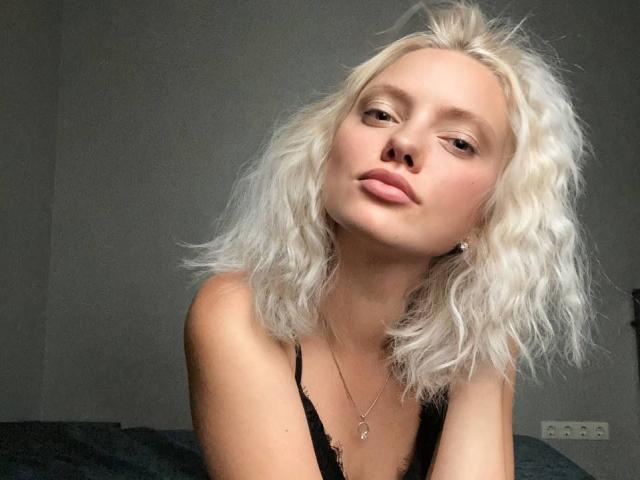 CrystalHope with messy white blonde hair, pouty pink lips, and a black lace top, looking slightly up and to the side, wearing a small necklace, in a soft light.