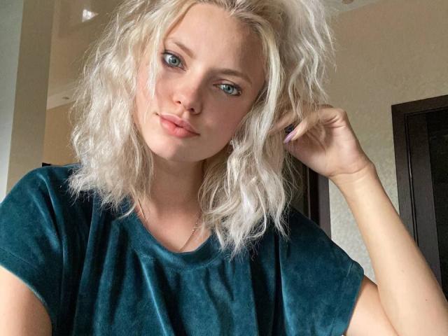 CrystalHope has wavy blonde hair, blue eyes, and a cute face with pink lips. She's wearing a green velvet shirt, touching her hair with a subtle smile.