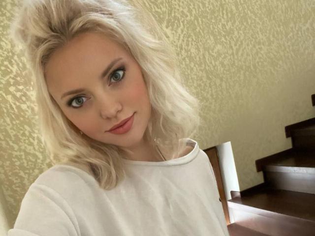 CrystalHope with blonde wavy hair, green eyes, and pink lipstick, wearing a white shirt, sitting at a wooden table, smiling softly, showing her cute face and subtle makeup.