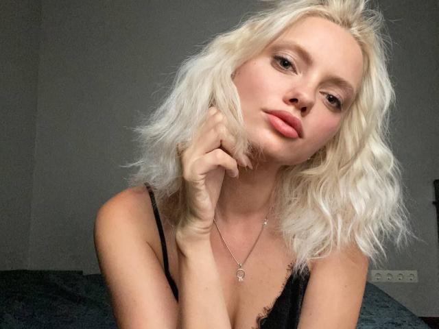 CrystalHope with platinum blonde, wavy hair, pink lips, and a slight smirk, wearing a black, lacy top, touching her cheek with her right hand, and a delicate necklace.