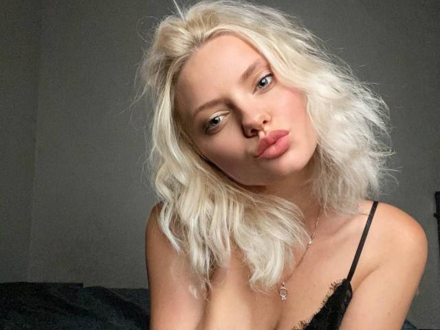 CrystalHope with pale blonde, wavy hair, pouty lips, and light skin, wearing a black spaghetti strap top, looking seductively at the camera. Her tits are slightly visible.