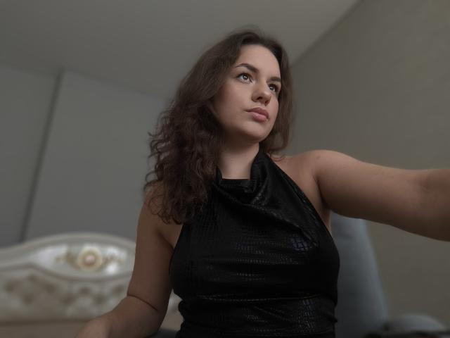 Dreia, avec ses cheveux bruns ondulés, porte un haut noir texturé sans manches et a une expression sérieuse. Elle tend le bras droit. Ses seins sont visibles, la lumière est douce..
