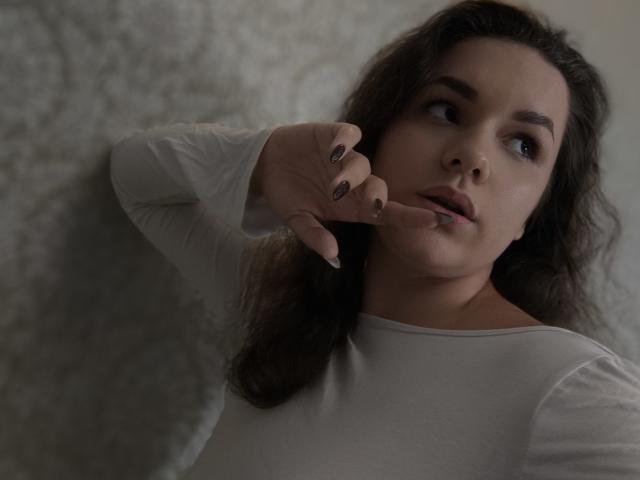 Dreia, avec ses cheveux bruns ondulés, un haut blanc à manches longues, un doigt près de sa bouche légèrement entrouverte, regardant sur le côté, un fond grisâtre, un vernis à ongles foncé, un éclairage doux, des lèvres légèrement entrouvertes..