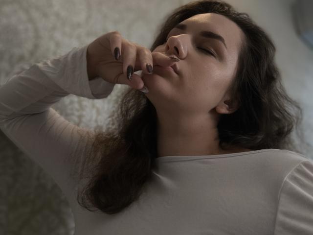 Dreia, les yeux fermés, le doigt sur les lèvres, portant une chemise blanche à manches longues, a les cheveux noirs et ondulés, et porte du vernis à ongles noir, et a l'air légèrement ennuyée..