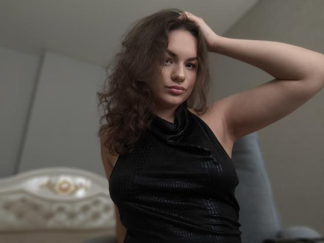 Dreia, avec ses cheveux bruns ondulés, porte un haut noir texturé sans manches. Sa main droite est dans ses cheveux, et elle regarde l'appareil photo avec un léger sourire, montrant sa poitrine..