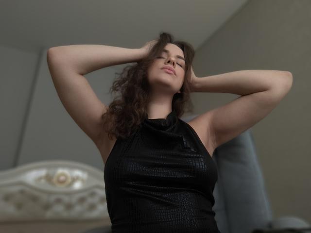 Dreia mit erhobenen Armen, Hände im braunen, lockigen Haar, trägt ein schwarzes, strukturiertes Top ohne Ärmel, leichtes Lächeln, weiches Licht, Oberkörper, Schlafzimmer, entspannt, selbstbewusst, attraktiv..