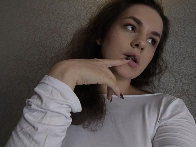 Dreia, avec ses cheveux bruns ondulés, porte une chemise blanche à manches longues, a un doigt dans la bouche, regarde sur le côté avec les lèvres légèrement entrouvertes, des ongles noirs et une expression neutre..