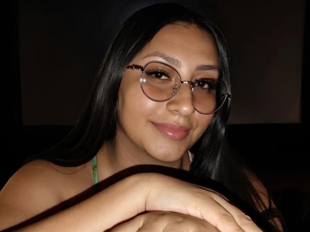 Catalina Rivero mit langem, schwarzem Haar, die eine Brille trägt und sanft lächelt, mit den Händen auf einer dunklen Oberfläche ruhend, ein grüner Riemen ist auf der Schulter sichtbar, dunkler Hintergrund, leicht glänzende, rosa Lippen..