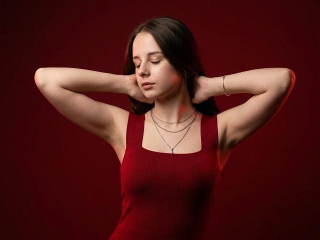 StacySpark, les bras levés, portant un débardeur rouge, montrant ses seins et ses aisselles, porte un bracelet et un collier, a les cheveux bruns, un fond rouge, les yeux fermés, une expression calme..