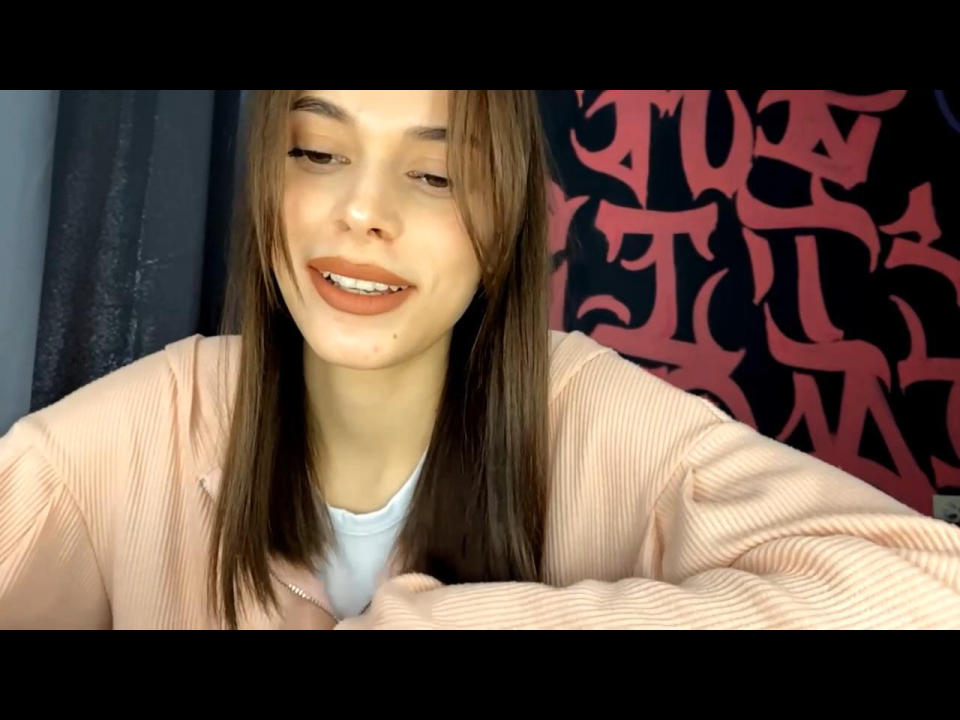 GwendolynRevalee with straight brown hair, light skin, and a cute smile, wearing a pink ribbed sweater and white shirt, showing a small beauty mark on her left cheek.
