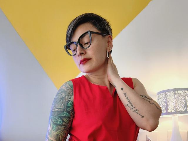 KatParadise with short black hair, glasses, red dress, tattoos on arms, holding neck, yellow and white wall, lamp with patterned shade, red lipstick, looking down, hand on neck.