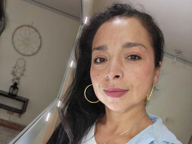 StangelarRivera has dark, wavy hair, brown eyes, and wears gold hoop earrings with a light blue shirt. Her face is slightly freckled with pink lipstick.