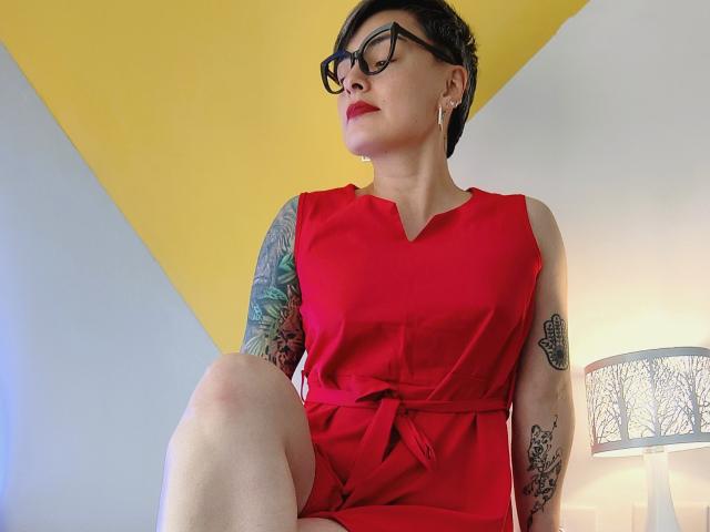 KatParadise with short black hair, glasses, red dress, and tattoos on arms, sitting with knee up, looking to the side, red lipstick, yellow and white wall background.