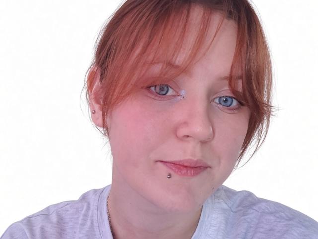 PatSanabria has red hair, blue eyes, and a pierced lower lip with a small black ball. She's wearing a light grey shirt, looking slightly tired.