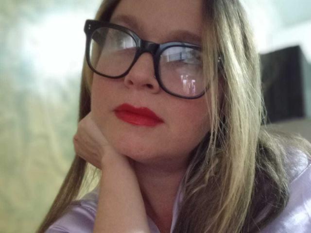 PamelaColin with glasses, red lipstick, and blonde hair, resting her chin on her hand, looking slightly up and to the side. She's wearing a shiny purple top.