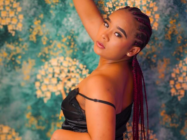 KailaniMoani with red braids, tan skin, and earrings, wearing a black strap dress, looking over her shoulder with a sultry expression, arm raised, showing her tits.