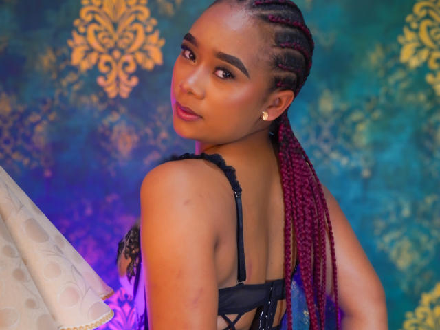 KailaniMoani with red braided hair, black lace bra, looking over shoulder, purple and blue lighting, small gold earring, soft smile, tan skin, curvy, cute, sexy.