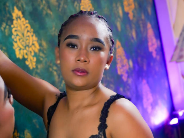 KailaniMoani with braided hair, dark eye makeup, glossy pink lips, black lace top, small earrings, looking at camera, arm raised, purple and green background, soft lighting.